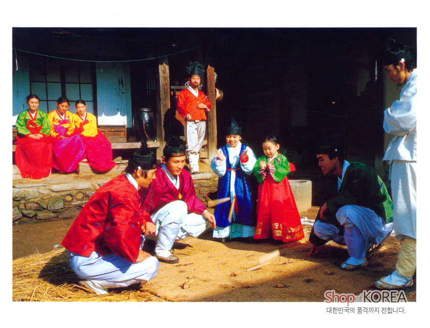 [텍스트: Shop of KOREA / 대한민국의 품격까지 전합니다.] 형형색색 비단 한복의 고운 색감과 전통 윷놀이가 어우러진 옛 기와집 마당 풍경 엽서 이미지