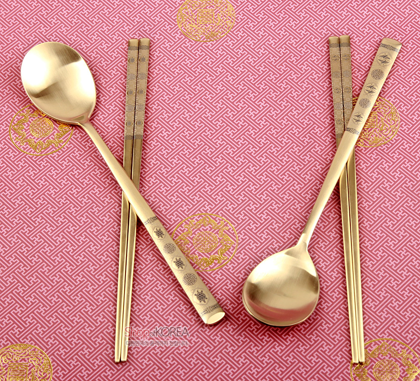 [텍스트: ShopofKOREA] 금빛 티타늄 수저·젓가락 2인 세트, 손잡이에 십장생과 기하학 문양 음각, 분홍 바탕 전통 문양 패턴 위 배치된 구성