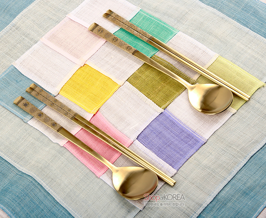 [텍스트: ShopofKOREA] 금빛 티타늄 소재 숟가락과 젓가락 2인 세트, 손잡이에 십장생과 기하학 문양 양각 장식, 다채로운 비단 조각보 위 연출 이미지
