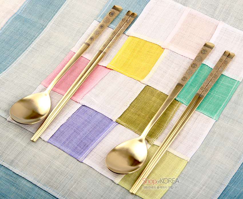 [텍스트: ShopofKOREA 대한민국의 혼을 세계까지 전합니다] 금빛 티타늄 수저와 젓가락 2인 세트, 손잡이에 십장생 문양과 기하학 문양 음각, 오방색 비단 조각보 위 연출 이미지