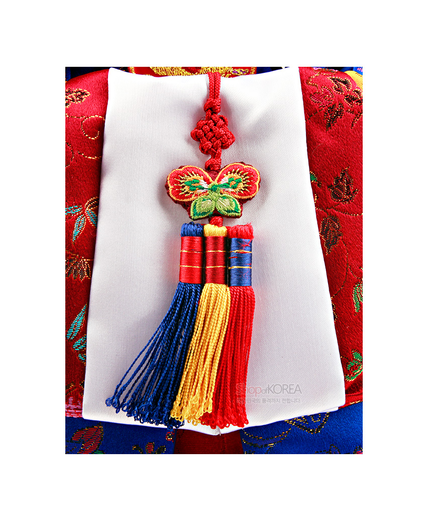 [텍스트: ShopofKOREA 한국의 문화를 전합니다] 화려한 비단 한복 위 흰 비단에 달린 나비 매듭과 오색 술 노리개 장식, 금사 꽃문양 자수 포인트의 전통 한복인형 디테일 묘사