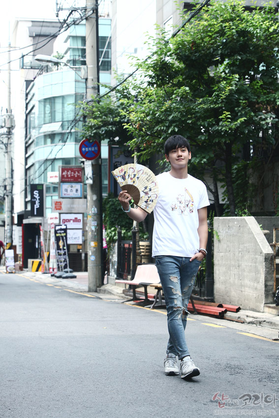 [텍스트: 샵오브코리아] 흰색 면 반팔 티셔츠에 다색 3D 입체발포 기법으로 표현된 풍물놀이 장단 인물 문양이 들어간 캐주얼 거리 풍경 이미지