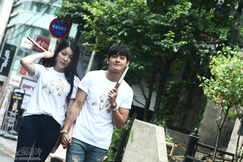 [텍스트: 샵오브코리아 대한민국 전통문화상품입니다] 순면 흰색 티셔츠에 다색 3D 입체발포 기법으로 표현된 풍물놀이 캐릭터 문양, 캐주얼한 거리 배경과 어우러진 연출 이미지