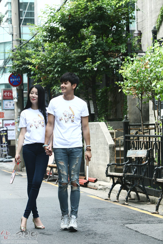 [텍스트: 샵오브코리아] 흰색 순면 반팔 티셔츠 위 다색 3D 입체발포 기법으로 표현한 풍물놀이 인물과 악기 문양, 캐주얼한 거리 배경 연출 이미지