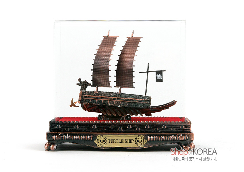 [텍스트: 龜, TURTLE SHIP, ShopKOREA 대한민국의 품격까지 전합니다.] 청동색 금속 거북선 모형과 붉은 벨벳 바닥, 장식문양 금속 받침대, 투명 아크릴 케이스가 조화를 이룬 전통 선박 장식품 모습