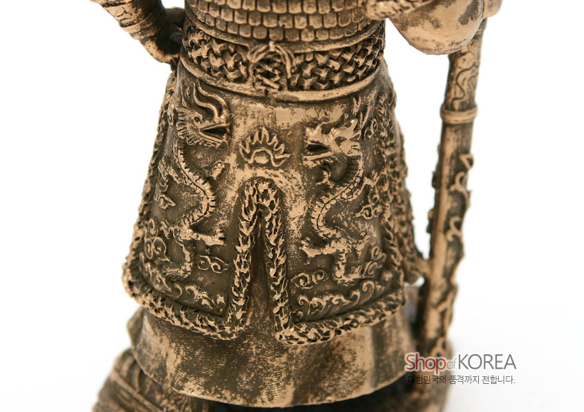 [텍스트: Shop of KOREA] 황동색 금속 재질의 이순신 장군 갑옷 부분 클로즈업, 앞치마에 입체적으로 새겨진 쌍룡 문양과 불꽃 장식, 비늘무늬 갑옷 질감이 돋보이는 조형미 표현
