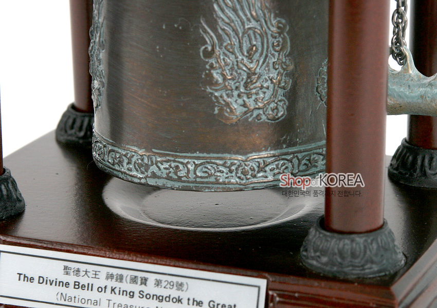 [텍스트: Shop of KOREA 대한민국의 품격까지 전합니다. / 聖德大王 神鐘(國寶 第29號) / The Divine Bell of King Songdok the Great] 청동색 동종 모형의 용무늬와 띠문양, 나무 기둥 받침대의 조합이 돋보이는 전통 장식품 모습