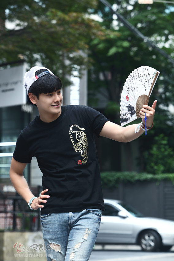 [텍스트: KOREA] 검정 면 티셔츠 위 금색 입체발포 호랑이 문양과 붉은 전각 스타일 낙관 포인트, 흰색 한글 서예 문양 부채를 든 연출 이미지