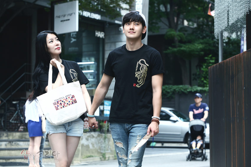 [텍스트: BEAUTIFUL KOREA] 검정 면 티셔츠에 금색 입체발포 호랑이 문양과 붉은 낙관 장식, 에코백 전면 직사각 패턴 인쇄가 돋보이는 거리 풍경 이미지 설명