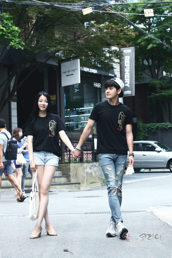 [텍스트: 샵오브코리아] 검정 면 티셔츠 위 금색 입체발포 호랑이 문양과 붉은 낙관 디테일이 도시 거리 배경 속에 어우러진 커플 연출 이미지