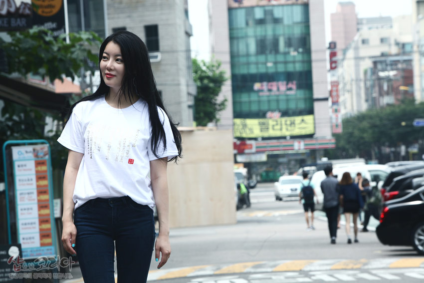 [텍스트: 샵오브코리아 대한민국 전통문화 공예의 멋이 깃들다] 흰색 면 티셔츠 위 금빛 입체발포 기법으로 표현된 훈민정음 한글·한자 문양과 붉은 낙관 포인트가 돋보이는 거리 연출 이미지