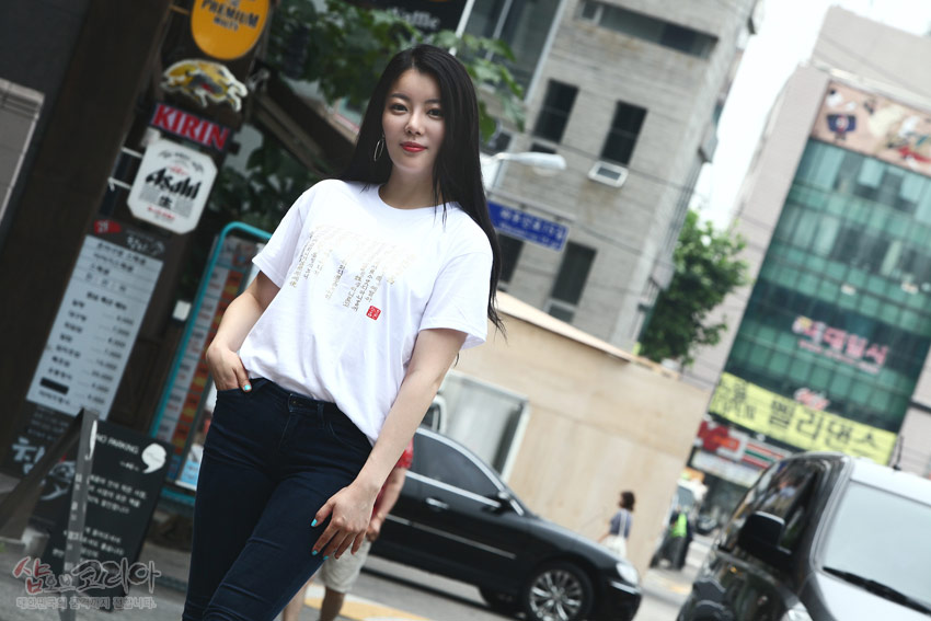 [텍스트: 훈민정음 서문 일부 금빛 한글·한자 혼합 문자] 흰색 면 반소매 위 금빛 입체발포 기법으로 세로 배열된 서체 문양 강조한 티셔츠 이미지