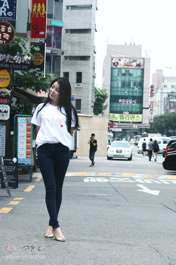 [텍스트: 샵코리아] 흰색 면 티셔츠에 금빛 입체발포 기법으로 훈민정음 한글·한자 서체 문양이 세로 배열된 디자인, 청바지와 조합된 거리 연출 이미지 설명