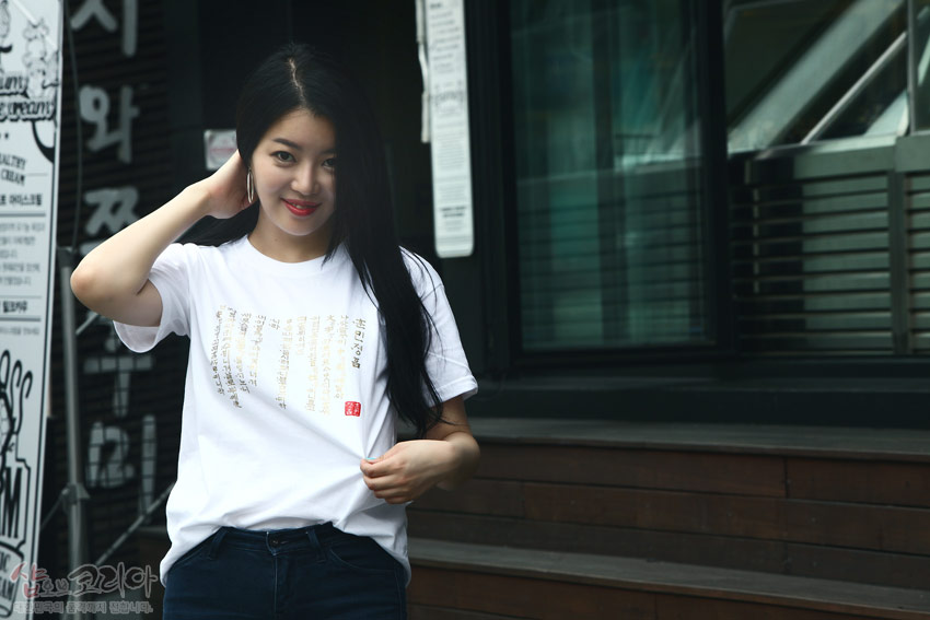 [텍스트: 훈민정음] 흰색 면 티셔츠 위 금빛 입체발포 기법으로 새긴 훈민정음 한글·한자 세로 배열 문양과 붉은 인장 포인트 디자인