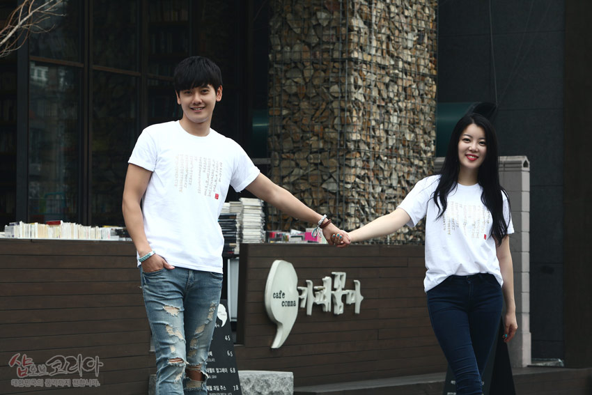 [텍스트: cafe comma] 흰색 면 티셔츠에 금빛 입체발포 기법으로 훈민정음 한글·한자 서체를 세로로 배치한 커플 연출 이미지