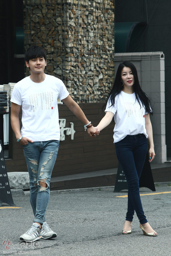 [텍스트: 훈민정음 한글·한자 문양] 흰색 면 티셔츠 위 금빛 입체발포 기법으로 세로 배열된 훈민정음 서체 문양이 강조된 커플 착용 연출 이미지