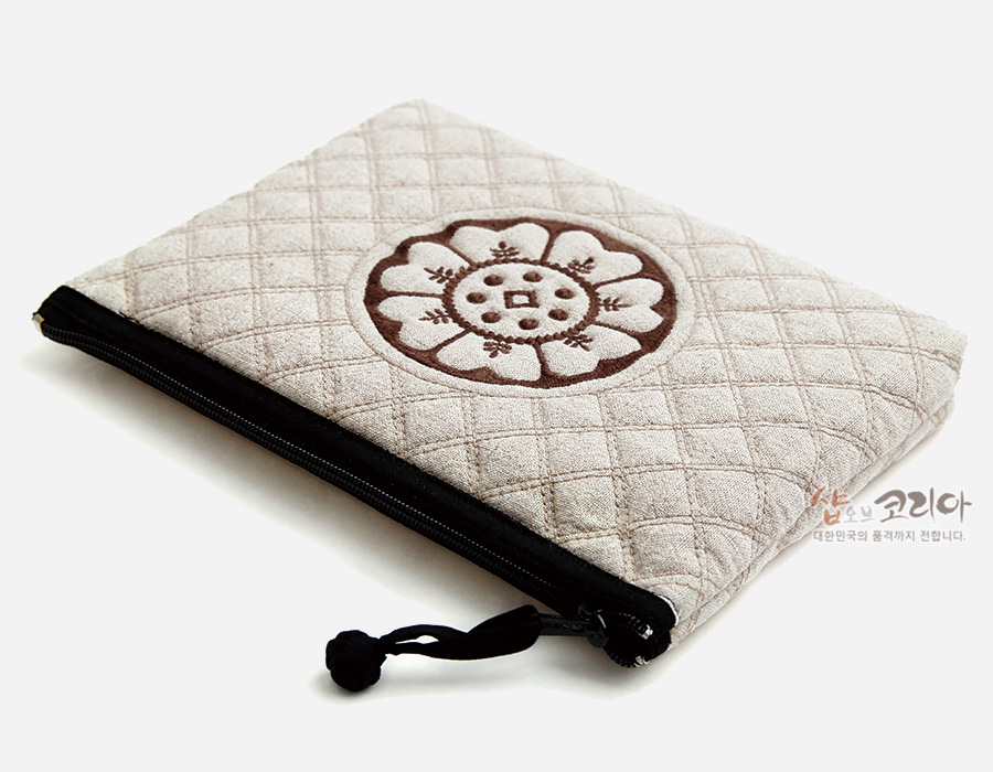 [텍스트: 샵오브코리아 대한민국의 품격까지 전합니다.] 연베이지색 누비 원단 위 갈색 백제 수막새 연꽃 문양 자수, 검정 지퍼와 끈 장식 사각 파우치 모습