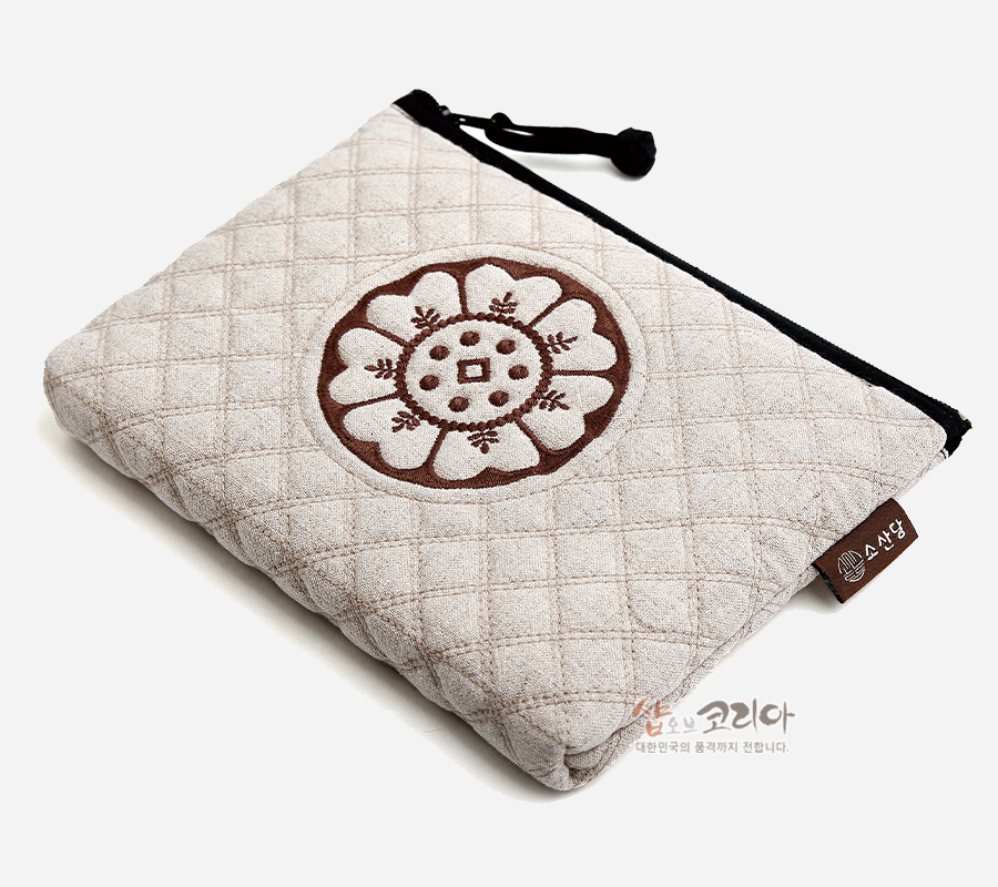 [텍스트: 샵오브코리아 소산당 대한민국의 품격까지 전합니다.] 베이지색 누빔 원단 위에 갈색 수막새 연꽃 문양 자수 장식, 검정 지퍼와 끈 디테일의 직사각 파우치 모습