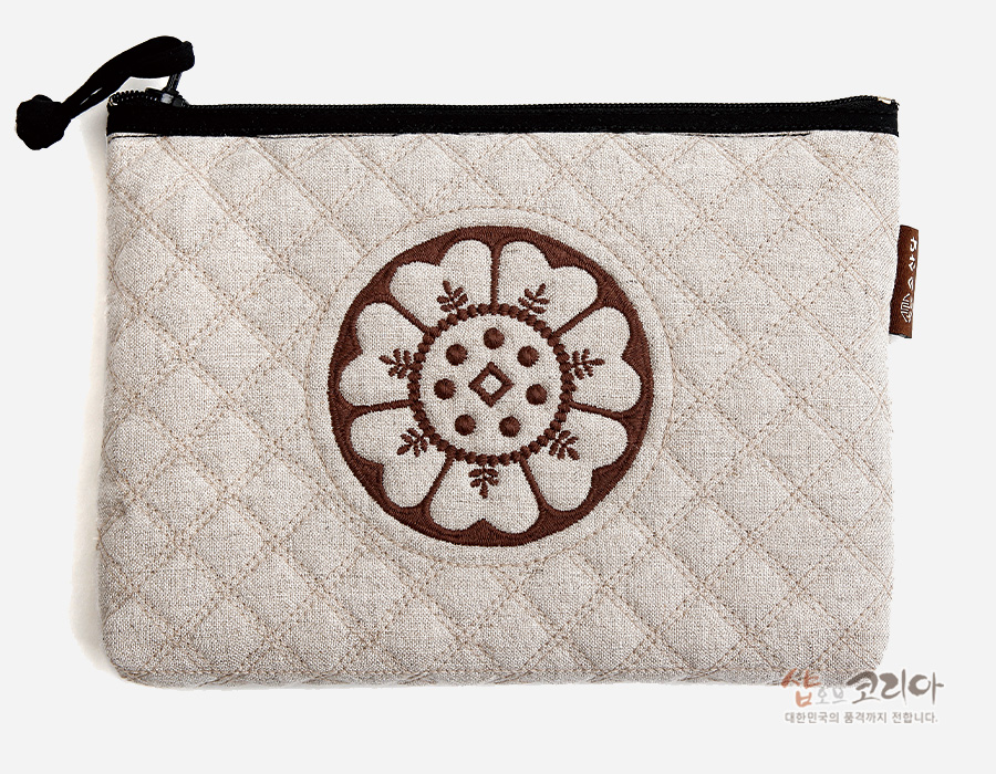 [텍스트: 샵오브코리아 대한민국의 품격까지 전합니다.] 베이지색 누빔 원단 위 갈색 백제 수막새 연꽃문양 자수 장식, 블랙 지퍼 마감 파우치 모습