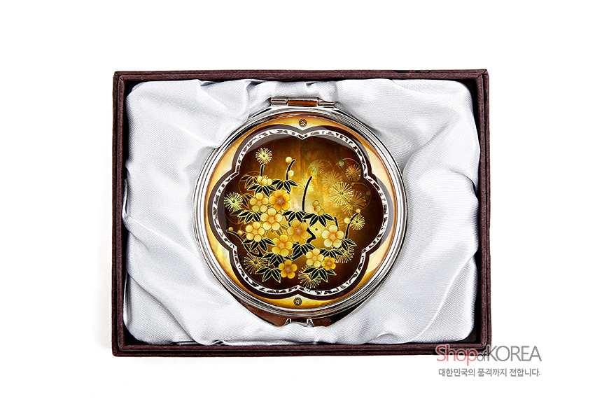 [텍스트: ShopKOREA 대한민국의 품격까지 전합니다.] 황색과 갈색 그러데이션 배경 위 자개 상감 기법으로 매화와 잎사귀 문양을 장식한 금속 원형 손거울 모습