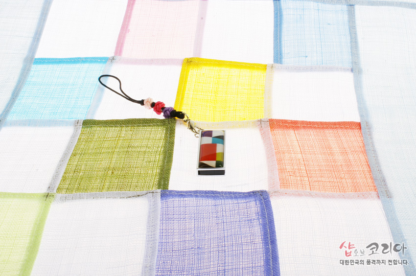 [텍스트: 샵오브코리아 대한민국의 품격까지 전합니다.] 금속 테두리 안에 빨강·노랑·초록·파랑 비단 조각보 문양을 넣은 직사각형 USB 메모리와 매듭 장식 스트랩, 반투명 색동 조각보 천 위 연출 이미지 설명