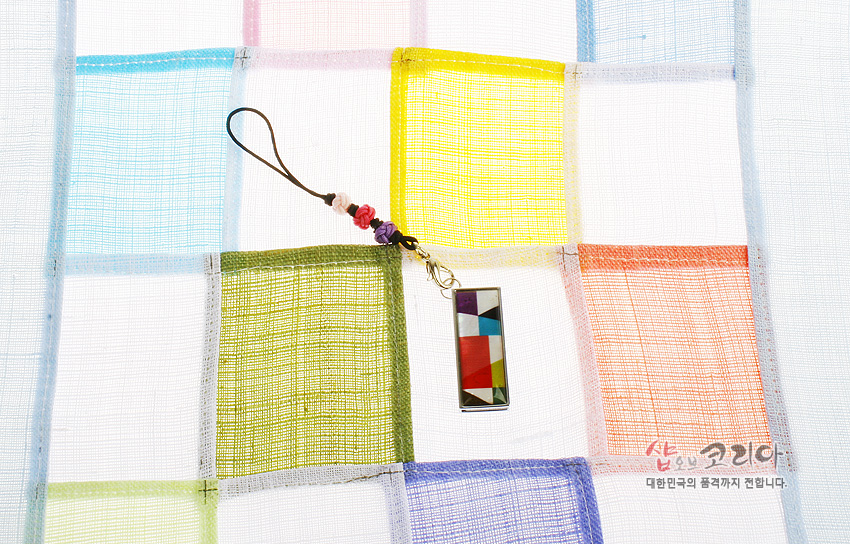 [텍스트: 샵오브코리아 대한민국의 품격까지 전합니다.] 금속 테두리 안에 붉은색·노란색·파란색 등 비단 조각보 패턴을 넣은 직사각형 USB 메모리와 매듭 장식 스트랩, 반투명 색동 조각보 배경 구성