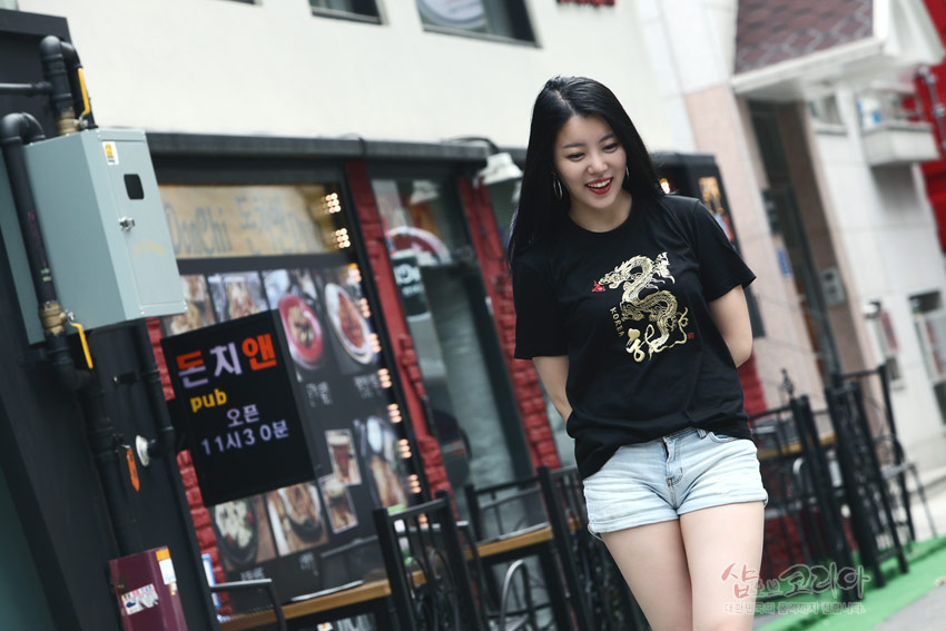 [텍스트: pub 오픈 11시 30분 KOREA] 검은 면 티셔츠 중앙 금색 3D 입체발포 용 문양과 붉은 포인트 장식, 거리 배경의 착용 연출 이미지 설명