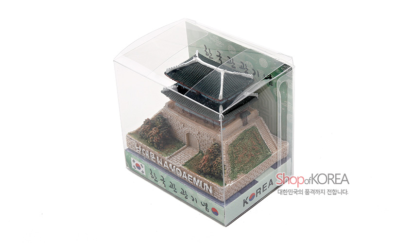 [텍스트: KOREA, NAMDAEMUN, Shop of KOREA, 대한민국의 품격까지 전합니다.] 투명 플라스틱 케이스 속 석고 재질 남대문 모형, 녹색 기와와 성곽, 수풀과 태극기 장식 구조물