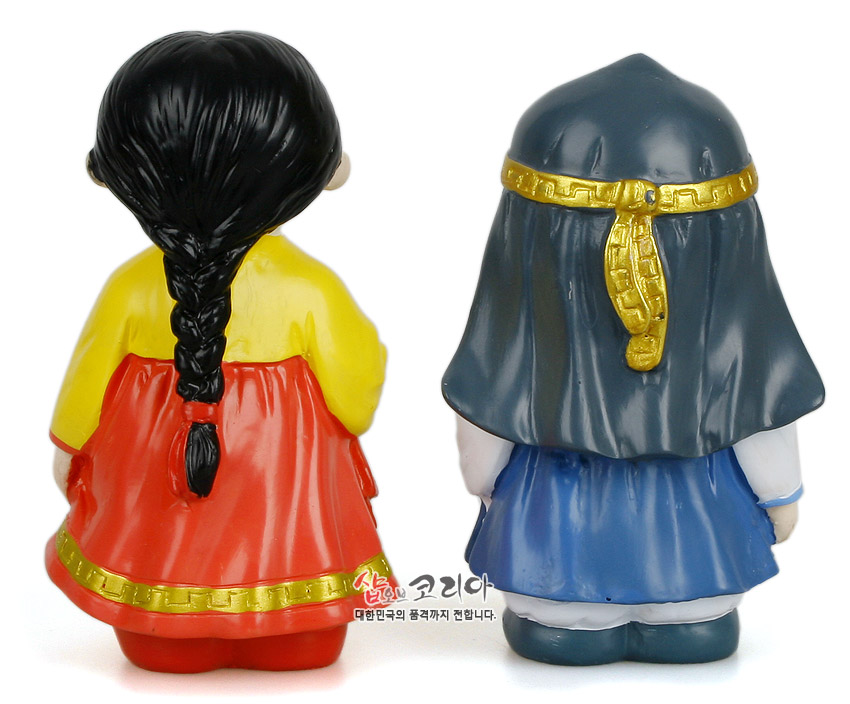 [텍스트: 샵오브코리아 대한민국의 품격까지 전합니다.] 채색된 플라스틱 재질의 춘향·몽룡 인형 뒷모습, 노랑 저고리와 붉은 치마·파랑 도포에 금색 띠 장식 대비가 돋보이는 구성