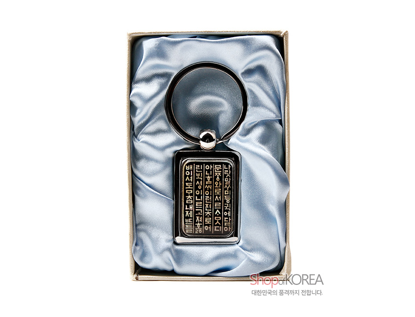 [텍스트: 나랏말쌈이 듕귁에 달아 문자와로 서르 사맛디 아니할쎄 이런 전차로 어린 백셩이 니르고져 홀 배 이셔도 마침내 제 뜨들 시러 펴디 못할 노미 하니라] 금속 테두리와 자개 상감 기법으로 훈민정음 글귀를 세로 배열한 사각 열쇠고리, 하늘색 실크 느낌 천에 올려진 구성