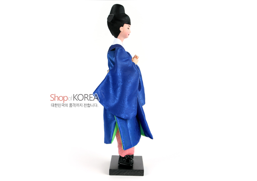 [텍스트: Shop of KOREA 대한민국의 품격까지 전합니다.] 청색 비단 도포에 은은한 꽃문양을 더한 혼례 신랑 인형, 분홍색 바지와 흑색 관모, 나무 받침대 조화 표현