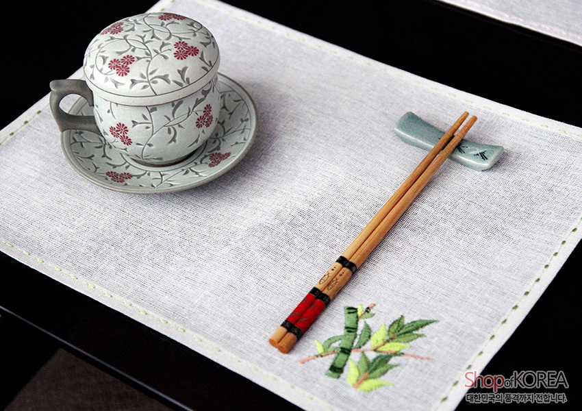 [텍스트: ShopofKOREA 대한민국의 품격까지 전합니다] 흰색 모시 식탁보 위 가장자리 연두색 스티치와 모서리 대나무 사군자 자수, 꽃무늬 도자기 찻잔과 젓가락 구성 이미지 설명