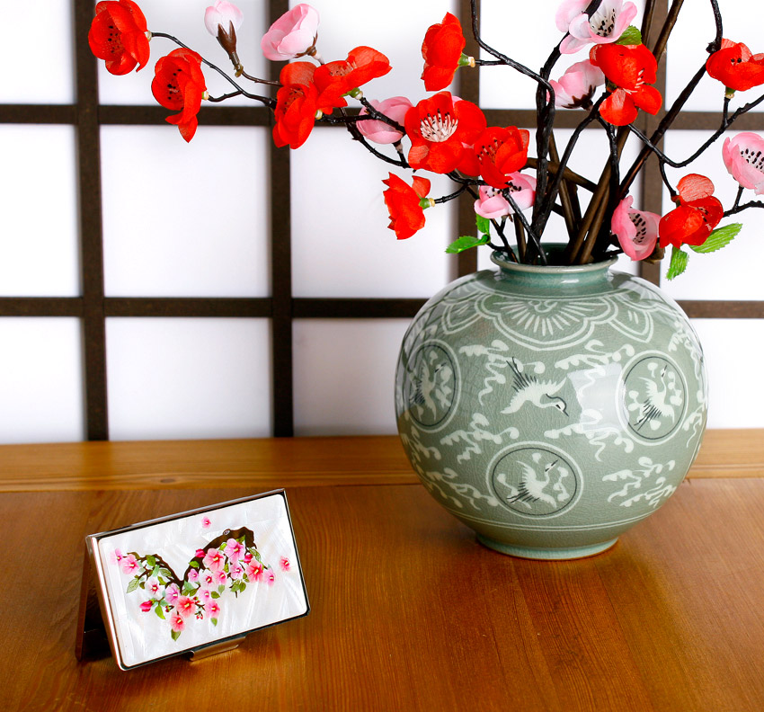 흰 자개 바탕 위 한반도 실루엣과 분홍 무궁화 문양을 상감 기법으로 장식한 금속 명함집, 청자 학무늬 꽃병과 조화가 어우러진 장식 연출 이미지