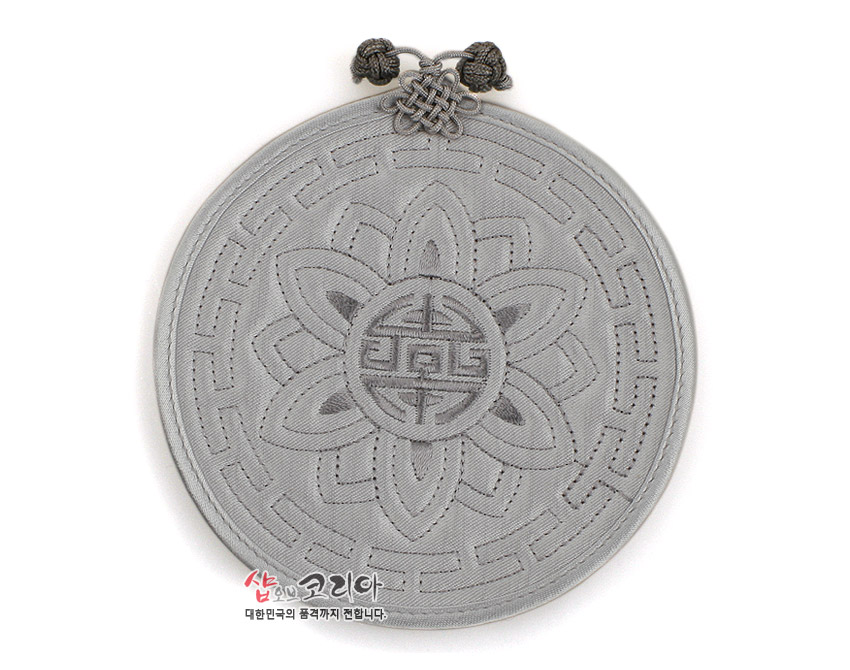 [텍스트: 샵오브코리아 대한민국의 품격까지 전합니다] 회색 패브릭 위 전통 누비 기법으로 연꽃과 수복문 문양을 새기고 상단에 매듭 장식을 더한 원형 컵받침 모습