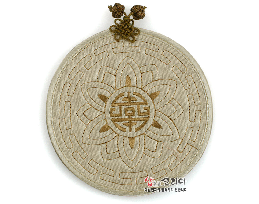 [텍스트: 샵오브코리아 대한민국의 품격까지 전합니다.] 베이지 톤 천 재질에 누비 기법으로 연꽃과 수복문 문양을 새기고 전통 매듭 장식을 더한 원형 컵받침형 패브릭 공예품