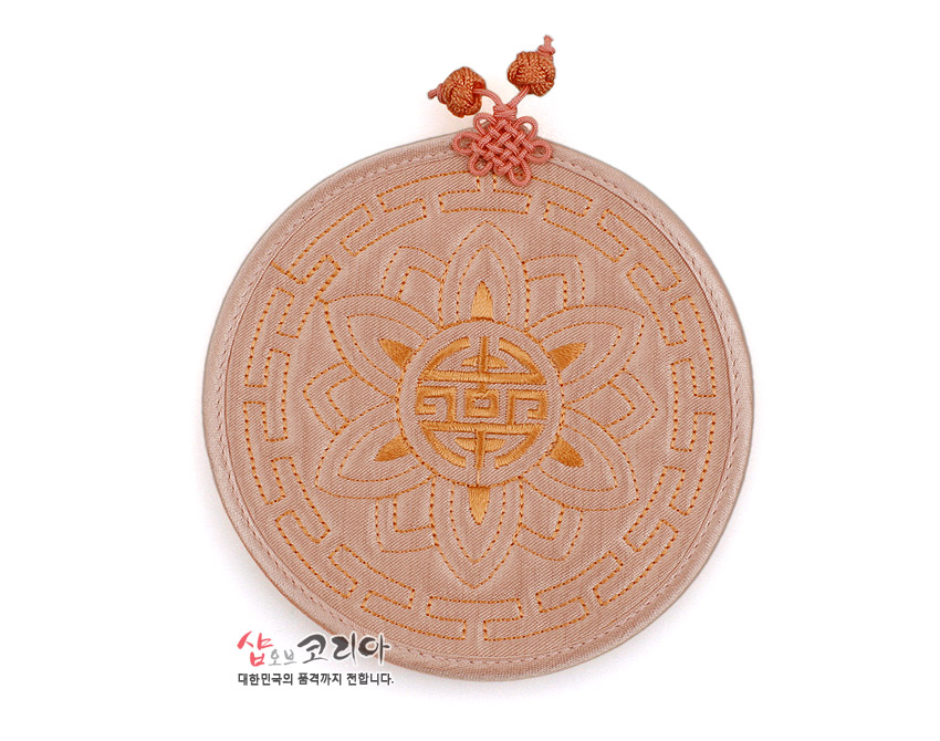 [텍스트: 샵오브코리아 대한민국의 품격까지 전합니다.] 파스텔 베이지 색 실크 누비 원형 컵받침, 연꽃과 수복문 자수 문양, 전통 매듭 장식 특징의 패브릭 공예품