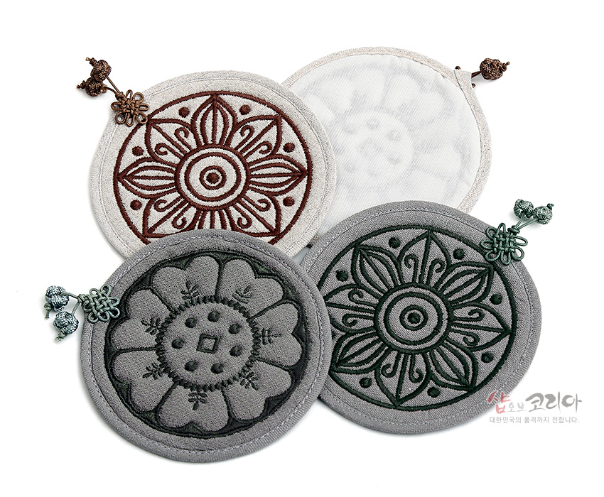 [텍스트: 샵오브코리아 대한민국의 품격까지 전합니다.] 회색·아이보리 천 위에 고구려·백제 수막새 연꽃문양을 누비 자수로 표현하고 색실 매듭 장식을 더한 원형 컵받침 구성