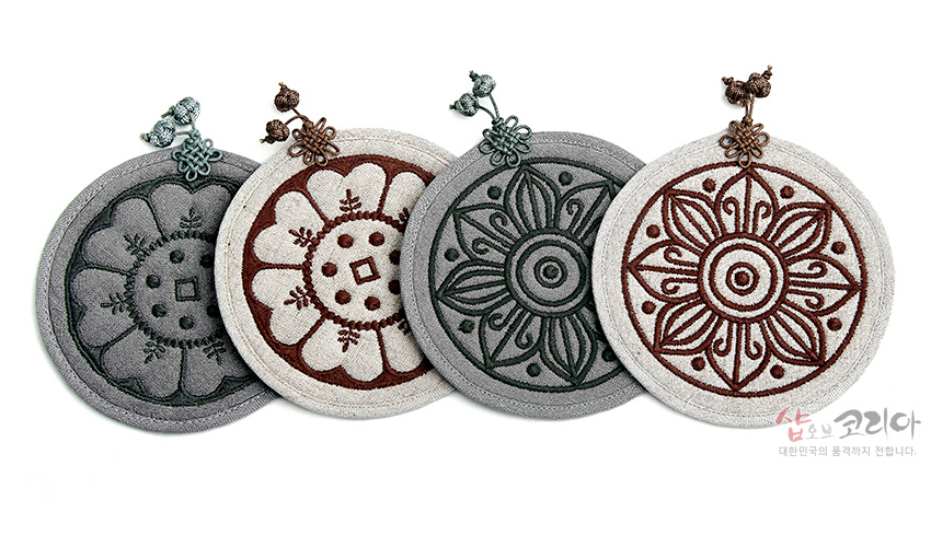 [텍스트: 샵오브코리아 대한민국의 품격까지 전합니다.] 회색 계열 천 소재에 고구려·백제 수막새 연꽃 문양을 자수로 놓고, 상단에 전통 매듭 장식을 단 원형 컵받침 2P 세트 이미지 설명