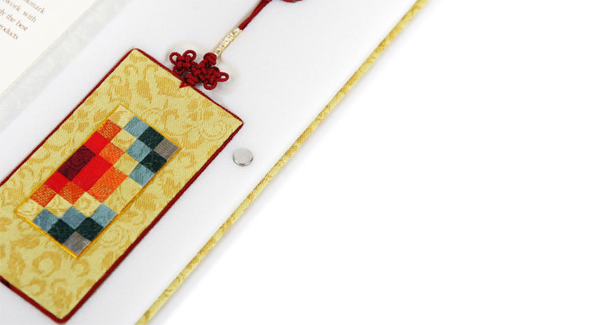 황색 비단 바탕에 전통 문양을 넣고 중앙에 다색 조각보 누비 패치워크를 더한 직사각형 책갈피와 붉은 전통 매듭 장식 구성 이미지