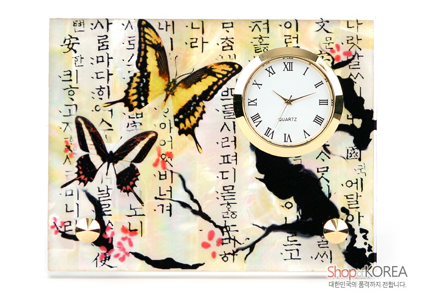 [텍스트: Shop of KOREA / 대한민국의 품격까지 전합니다.] 자개 바탕 위에 옻칠 기법으로 매화 가지와 분홍 매화꽃, 노랑·갈색 나비 문양을 넣고 금속 테두리 로마 숫자 시계를 더한 직사각형 탁상용 시계 이미지 설명