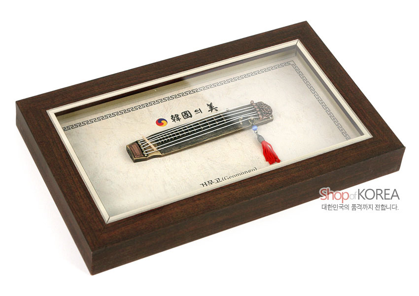 [텍스트: 韓國의 美, 거문고(Geomungo), Shop of KOREA, 대한민국의 품격까지 전합니다.] 짙은 갈색 나무 액자 속 금속 미니어처 거문고와 태극 문양, 붉은 술 장식이 어우러진 전통 악기 장식 이미지