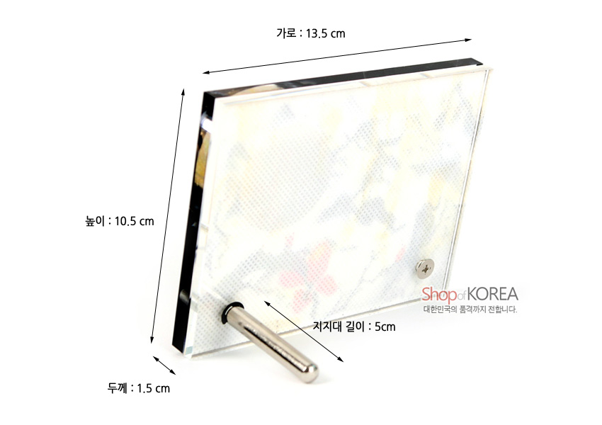 [텍스트: 가로 : 13.5 cm, 높이 : 10.5 cm, 두께 : 1.5 cm, 지지대 길이 : 5cm, Shop of KOREA 대한민국의 품격까지 전합니다.] 연한 산수도와 붉은 꽃문 양이 비치는 자개 배경, 금속 프레임과 원통형 금속 지지대로 구성된 직사각형 탁상시계 뒷면 모습