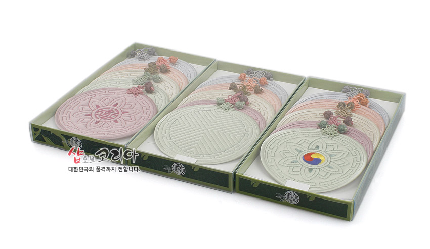 [텍스트: 샵오브코리아 대한민국의 품격까지 전합니다.] 파스텔 톤 원형 천 소재에 전통 누비 기법으로 기하학·연꽃·태극 문양을 넣고 배씨머리 매듭 장식을 더한 컵받침 세트 구성