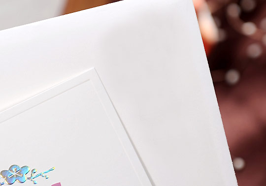 부드러운 백색 종이 재질의 연하장 일부 클로즈업, 가장자리 엠보싱 처리와 홀로그램 느낌의 꽃 문양 포인트가 돋보이는 미니멀 디자인 연하장 이미지