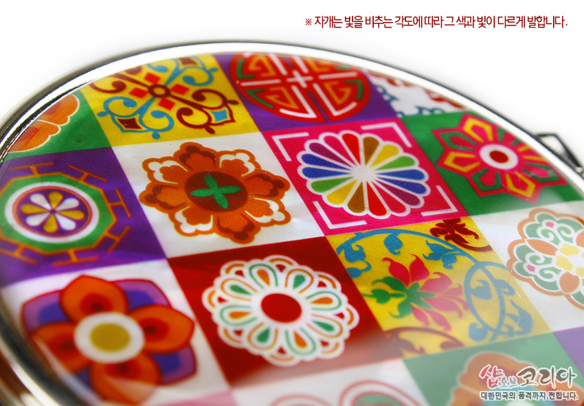 [텍스트: ※ 자개는 빛을 비추는 각도에 따라 그 색과 빛이 다르게 발합니다.] 금속 테두리 안 자개 표면 위에 다채로운 색상의 전통 꽃문양과 기하학 무늬가 격자 형태로 배열된 손거울 장식 이미지
