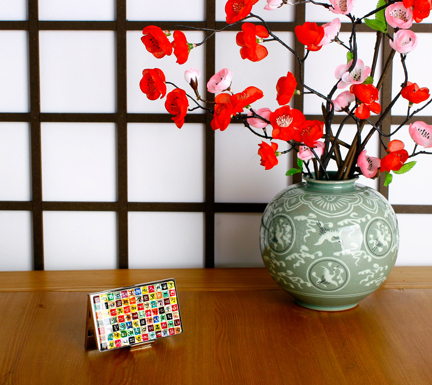 [텍스트: 한글 자모] 자개 바탕 위 다채로운 색상의 정사각형 칸마다 한글 자모를 배열한 금속 명함집과 청자 문양 화병의 조화로운 연출 이미지