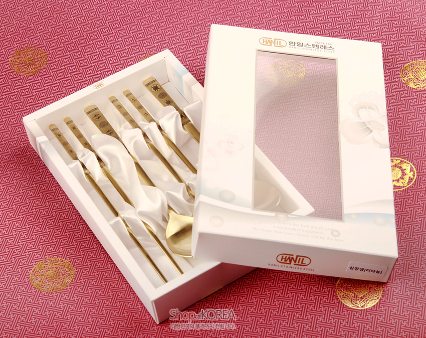 [텍스트: HWIL STAINLESS STEEL, ShopKOREA, 십장생(티타늄)] 분홍 전통 기하학 문양 바탕 위 흰 케이스에 담긴 금빛 티타늄 수저, 손잡이에 십장생과 한자 문양이 새겨진 디자인