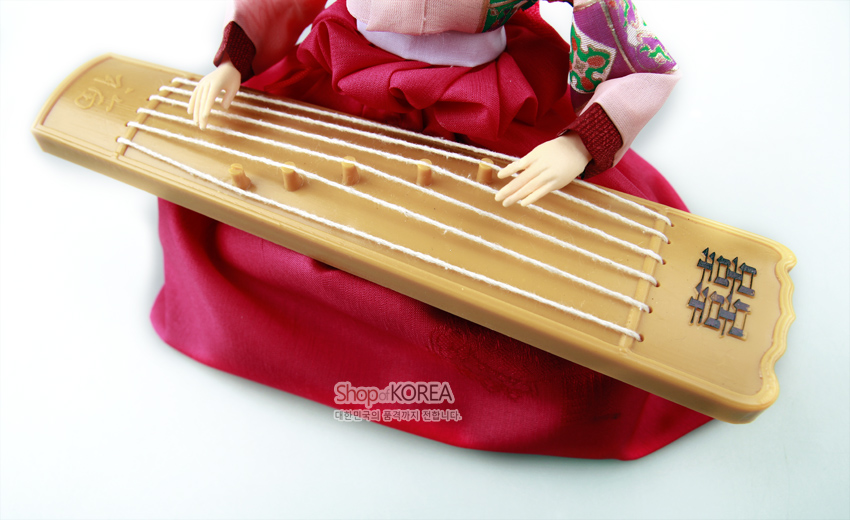 [텍스트: Shop of KOREA 대한민국의 전통미가 살아납니다] 분홍 비단 치마와 색동 저고리를 입은 인형이 연주하는 나무 가야금과 흰 줄, 양각 문양 장식 모습