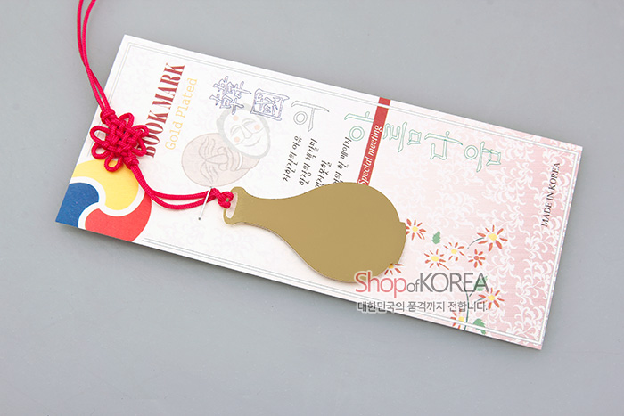 [텍스트: BOOKMARK, Gold Plated, Shop of KOREA, 대한민국의 품격까지 전합니다] 금장 금속 청자 주병 실루엣 책갈피와 붉은 전통 매듭끈, 연분홍 꽃무늬 배경 카드 조화 이미지