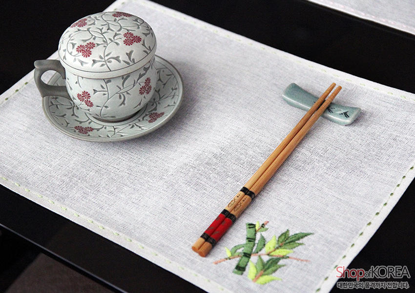 [텍스트: Shop of KOREA 대한민국의 품격까지 전합니다] 흰색 모시 식탁보 위 연두색 가장자리 스티치, 대나무 자수 문양과 전통 문양 찻잔, 나무 젓가락과 도자기 받침 구성 이미지